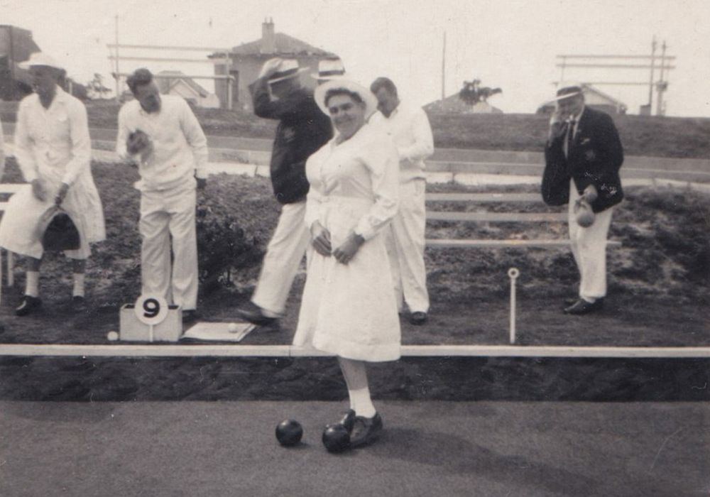 Black and white photo: a woman bowler in whites delivers a bowl as others watch