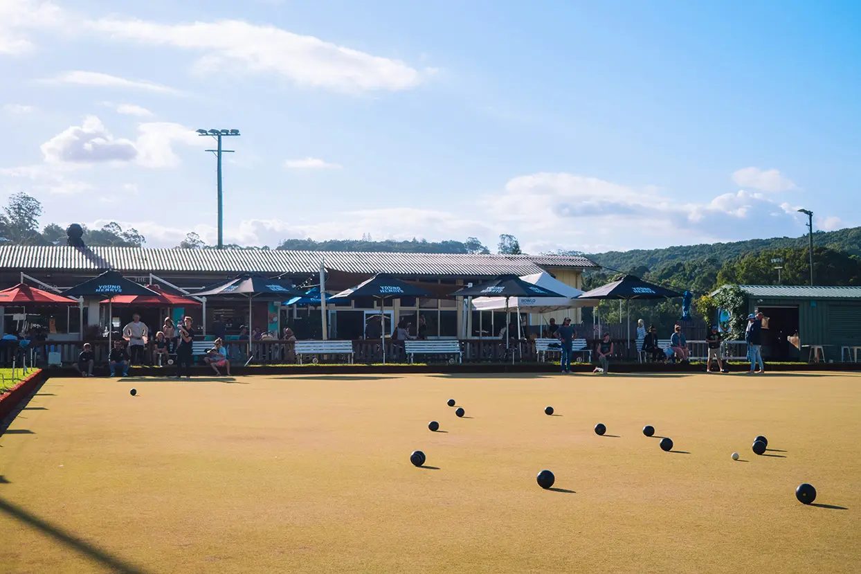 Bowls scattered across the green on a sunny afternoon, with the clubhouse and umbrellas behind
