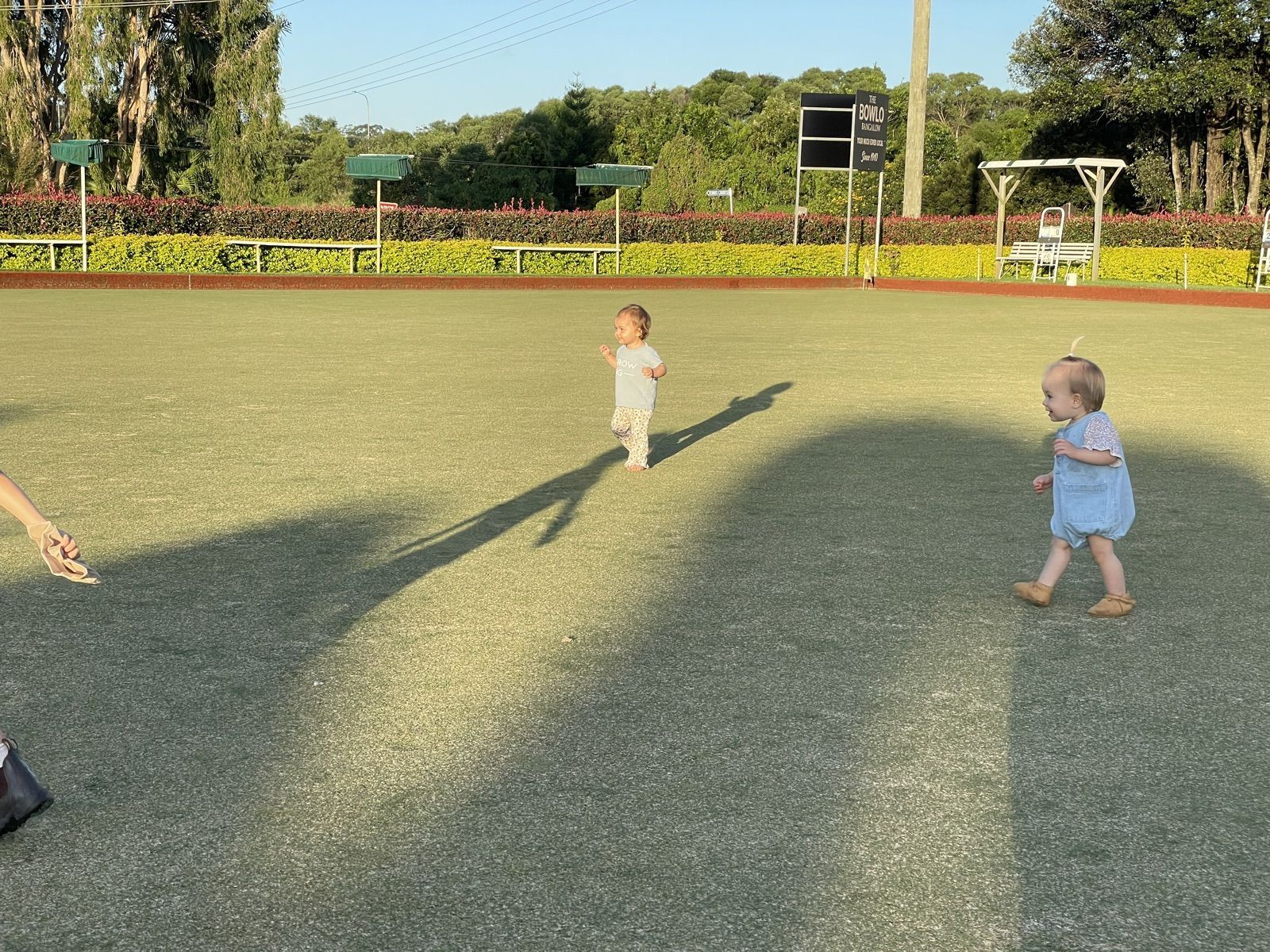 Two toddlers walking barefoot across the Bowlo's bowling green in the late afternoon sun