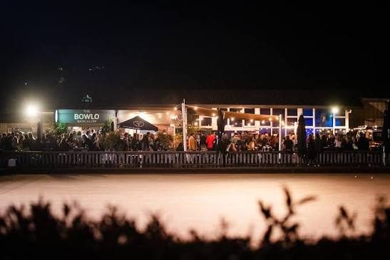 The Bowlo's verandah lit up at night and full of people, seen across the dark green